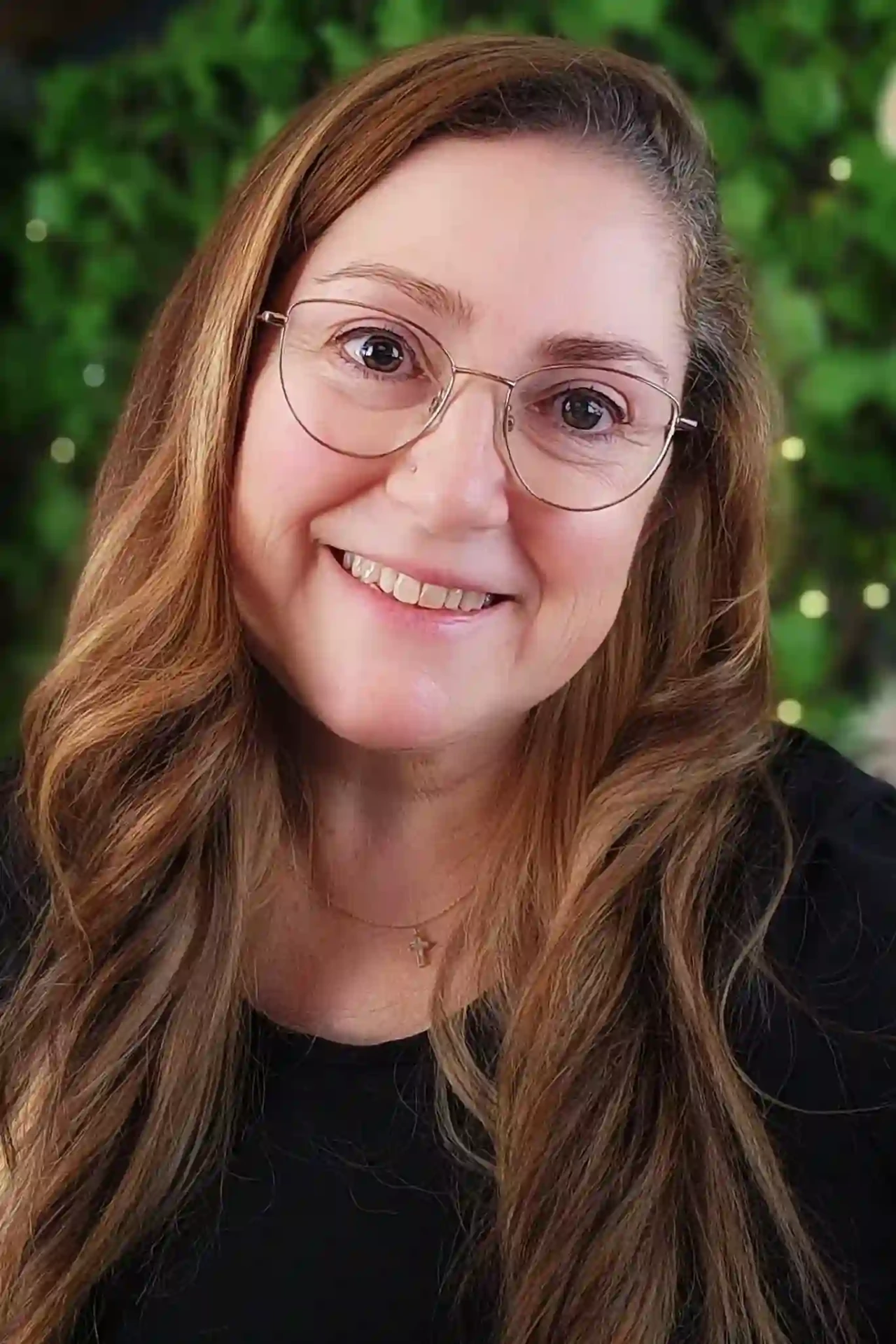 Portrait photograph of Dr Fiona Girkin smiling, wearing glasses and a black top, against a green foliage background with soft fairy lights. She is an Australian consultant, researcher and advocate for evidence-based conversations on gender, trauma and justice.